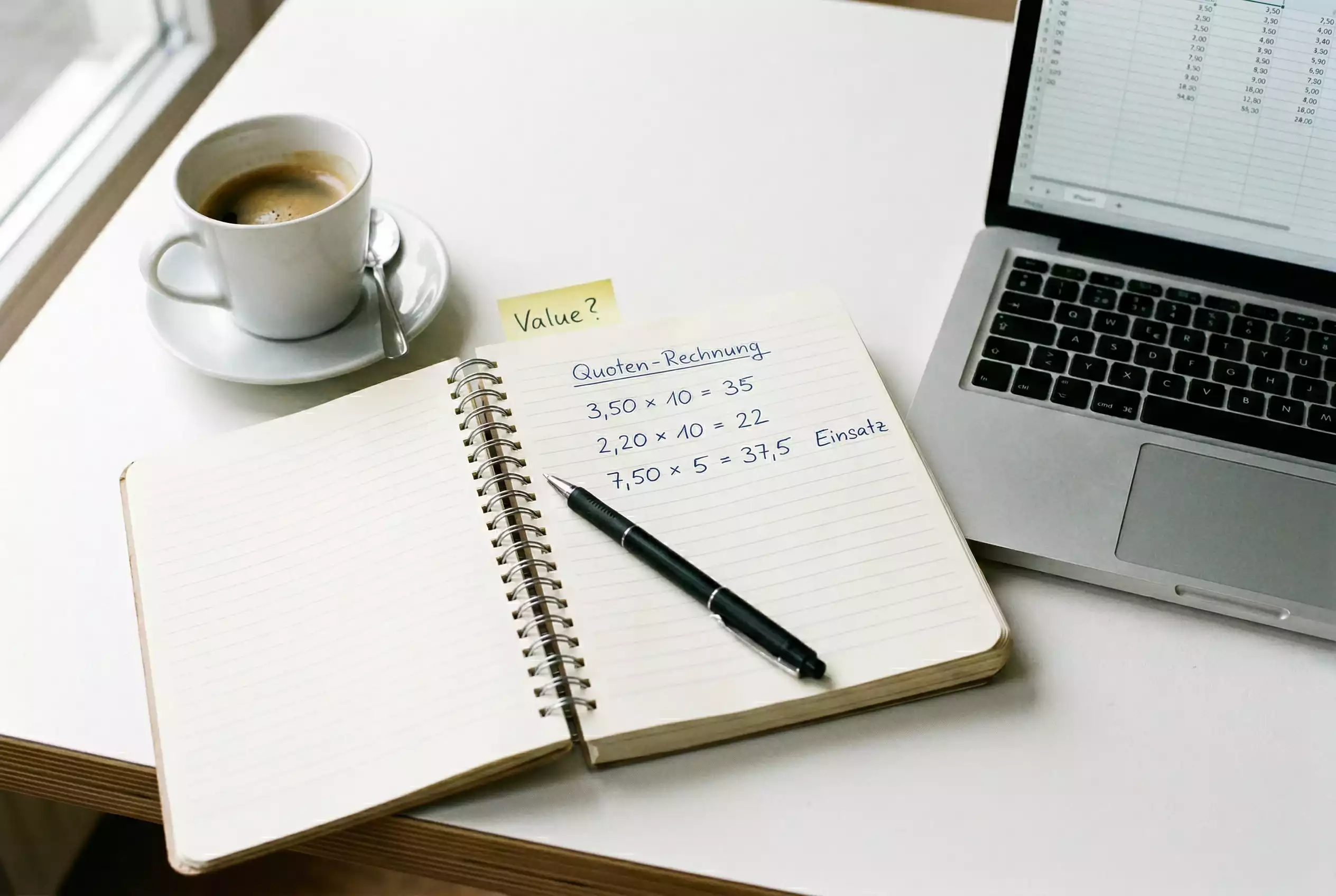 Nahaufnahme eines Notizblocks mit handgeschriebenen Quotenberechnungen neben einem Laptop